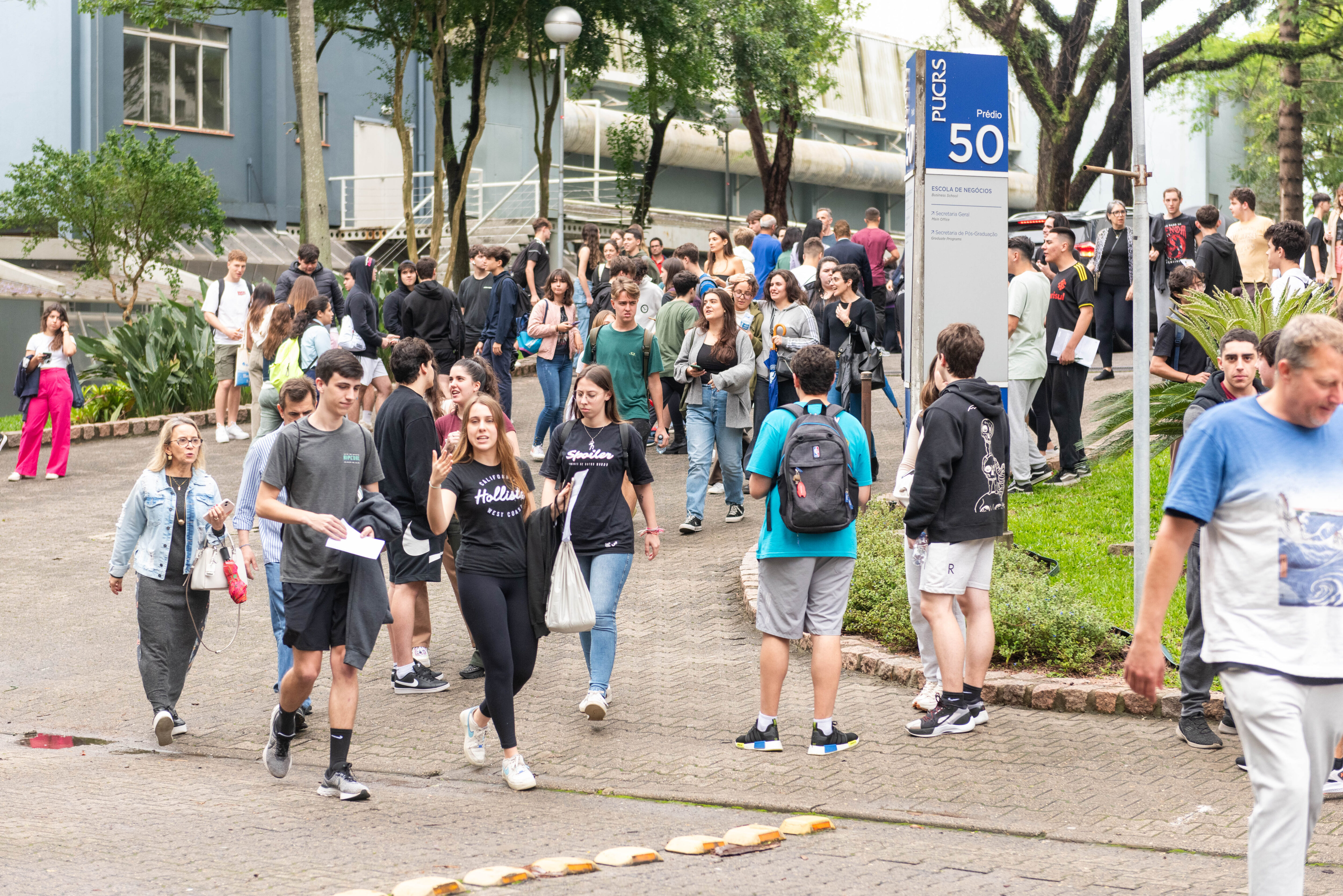 Foto tirada em área externa do campus da PUCRS, com muitas árvores ao fundo e prédios em tons de azul. A imagem mostra uma movimentação intensa de pessoas, a maioria jovens estudantes, caminhando em diferentes direções. No centro da imagem, há uma placa azul e branca com a inscrição “PUCRS – Prédio 50 – Escola de Negócios”. Muitas pessoas estão conversando, algumas segurando papéis e mochilas, vestindo roupas casuais como camisetas, calças jeans, shorts e tênis. À esquerda, uma mulher mais velha, de óculos e cabelos loiros, também caminha pelo local. Ao fundo, observa-se uma escada metálica, vegetação e parte de um tubo de ventilação industrial que atravessa o prédio. O ambiente é arborizado e transmite um clima de integração universitária.