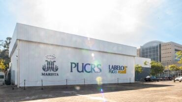 Fachada de um prédio branco com os logotipos do Marista, PUCRS e LABELO PUCRS estampados na lateral. O céu está claro e ensolarado, com alguns reflexos de luz na imagem. Ao fundo, há um edifício moderno de vidro e concreto, além de árvores e carros estacionados.