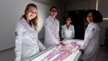 Quatro pessoas vestindo jalecos brancos estão reunidas ao redor de uma mesa interativa de anatomia digital em uma sala de aula. Duas mulheres e um homem jovens estão sorrindo, enquanto uma mulher mais velha, que parece ser professora, também sorri para a câmera. O ambiente tem iluminação suave, com a mesa digital destacando a imagem detalhada de um corpo humano exibido na tela. Os jalecos possuem logotipos da PUCRS e do Hospital São Lucas, indicando uma conexão com a área da saúde e educação médica.