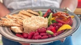 Uma pessoa segura um prato redondo com uma refeição composta por tiras de filé de frango grelhado à esquerda, um montinho de arroz integral ao centro, feijão-vermelho ao redor na parte inferior, e, à direita, legumes grelhados — pimentões vermelho, amarelo e laranja, brócolis e fatias de cenoura. A pessoa veste uma camisa clara e apoia o prato com as duas mãos, ligeiramente desfocadas ao fundo.