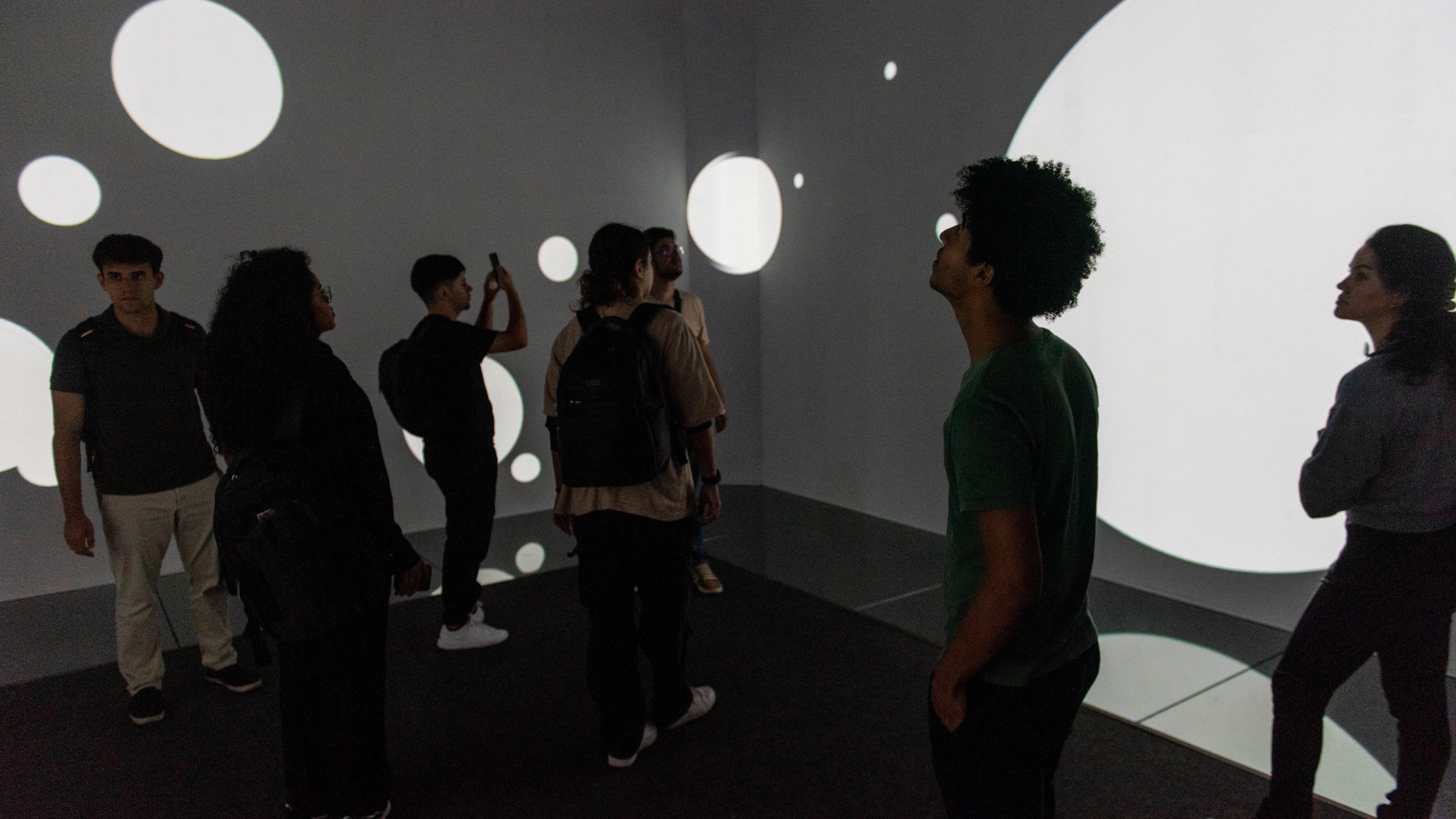 Grupo de pessoas observa projeções de círculos brancos em diferentes tamanhos sobre as paredes e o chão de uma sala escura, criando um ambiente imersivo. Alguns olham atentamente para as imagens, enquanto outros registram o momento com o celular. Todos estão em pé, com mochilas nas costas, explorando a experiência sensorial.