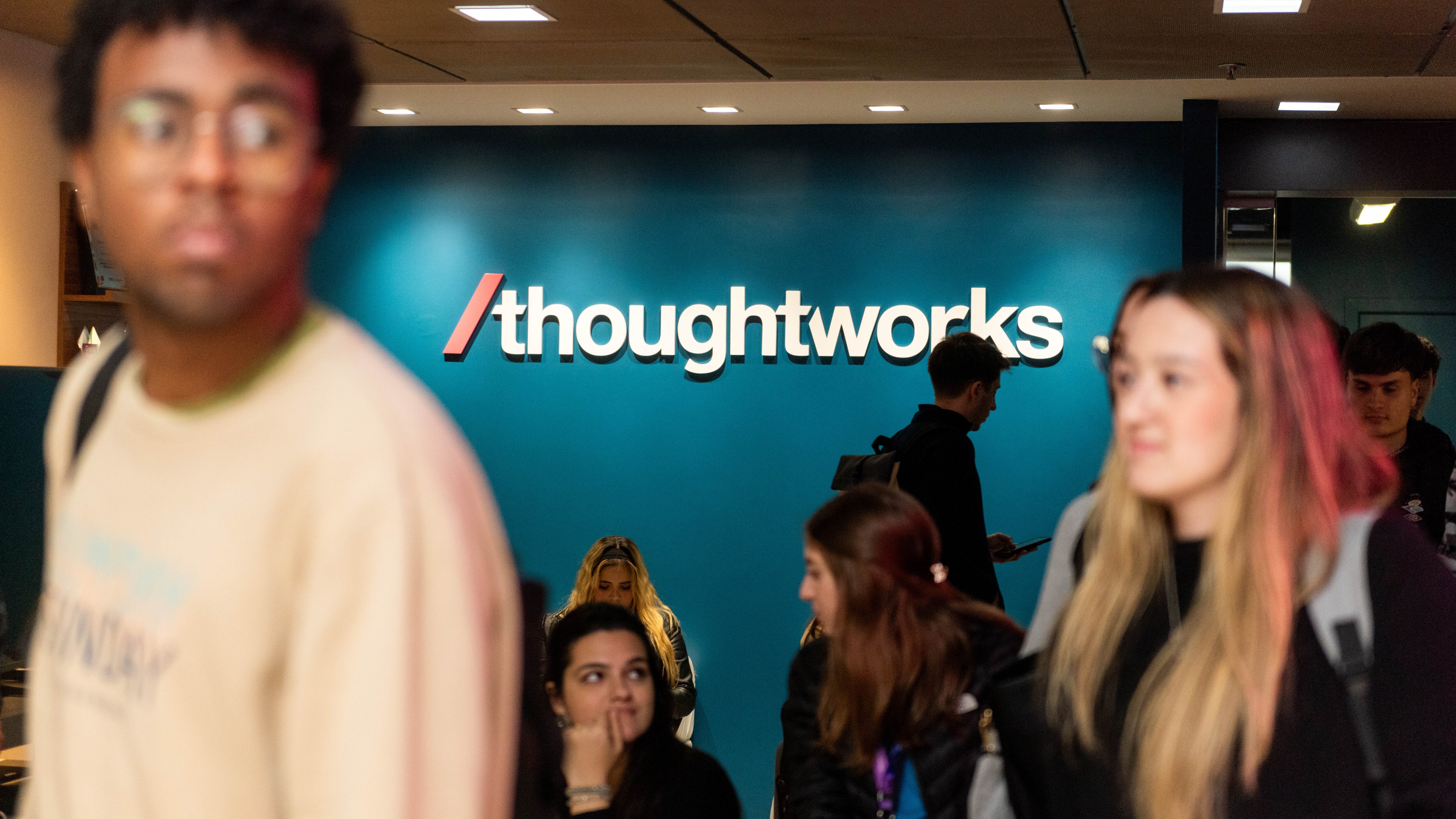 Pessoas circulam e conversam em frente a uma parede azul com o logo da empresa Thoughtworks em destaque. Algumas estão sentadas, outras em pé, utilizando mochilas e celulares. O ambiente tem iluminação clara e transmite um clima de interação e movimento.