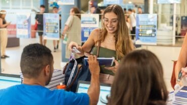Jovem sorridente entrega um crachá para um atendente durante um evento em ambiente universitário. Ela segura materiais como cadernos e uma bolsa, enquanto o atendente, que está de camiseta azul, faz o registro em um notebook. Ao fundo, há outras pessoas circulando e cartazes de divulgação com informações sobre a universidade.