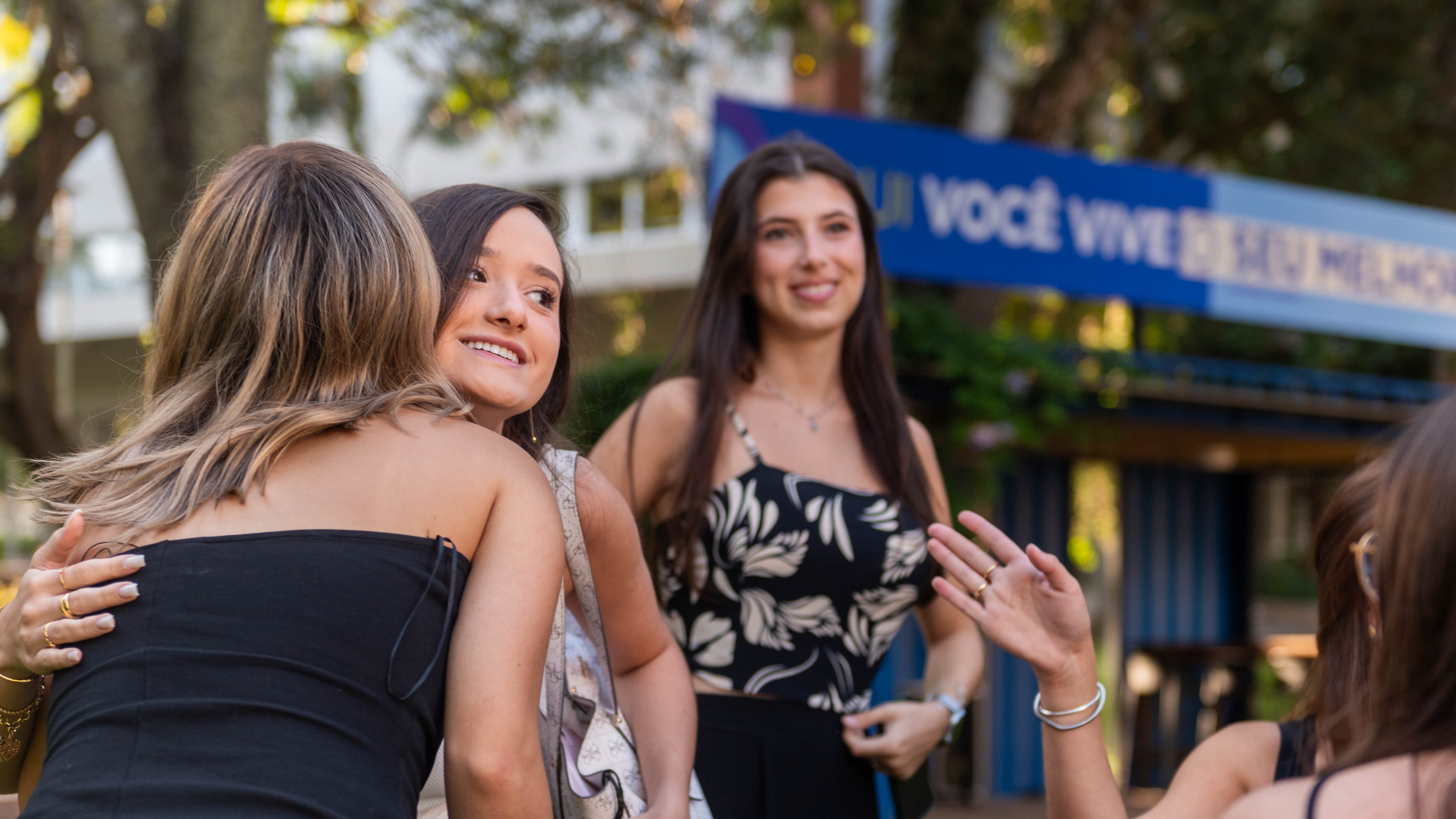Grupo de jovens se cumprimenta com abraços e sorrisos em um ambiente ao ar livre, com árvores ao fundo e uma placa azul desfocada que traz a frase ‘Aqui você vive o seu mundo’. O clima é de acolhimento, descontração e reencontro, possivelmente no primeiro dia de aula ou em um evento universitário.”