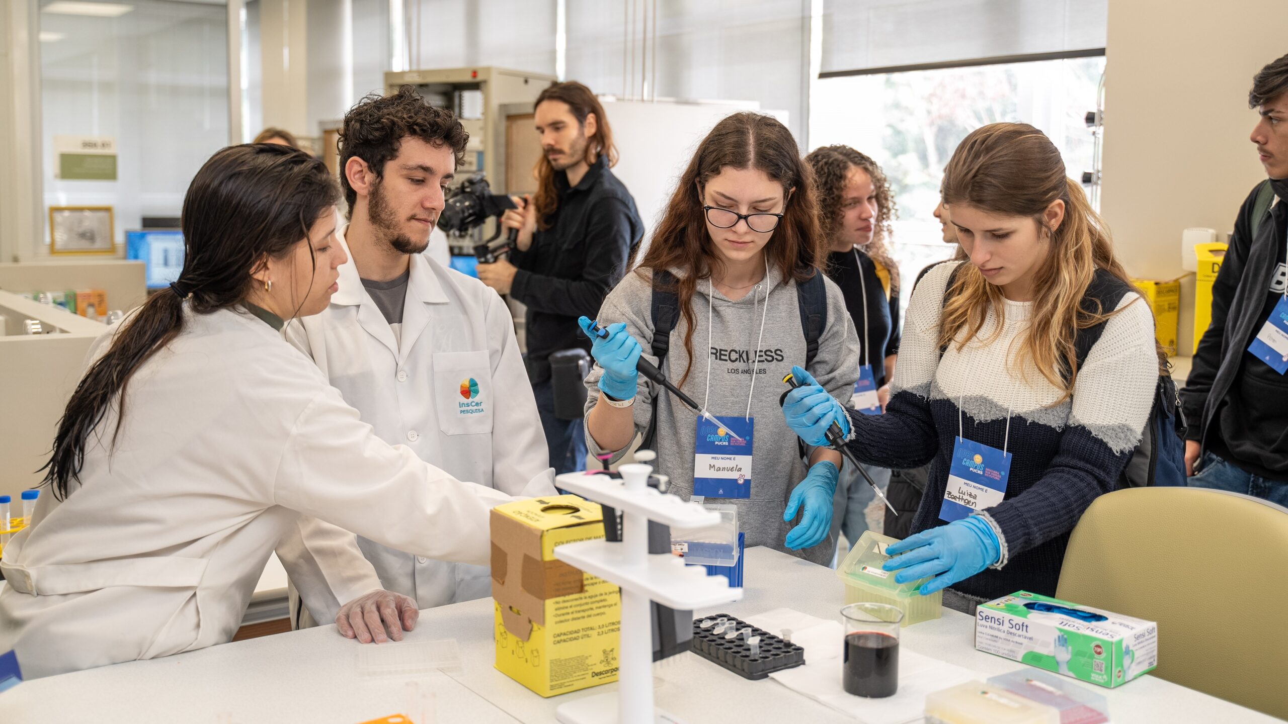 Grupo de estudantes participa de uma atividade prática em um laboratório de pesquisa. Duas alunas manuseiam micropipetas, usando luvas azuis, enquanto recebem orientação de uma pesquisadora e um pesquisador de jaleco do Instituto do Cérebro (InsCer). Ao fundo, outros participantes observam. O ambiente é moderno, bem iluminado e equipado com materiais científicos.

