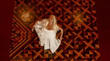Fafá de Belém, blonde with long, wavy hair, wears an elegant white dress with a lace cape, smiling and looking up. She is positioned in the center of a wooden floor with rich geometric designs in shades of brown, red and gold, which form a visually striking mosaic.