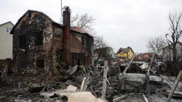 Destroços carbonizados de uma aeronave não identificada espalham-se por uma área residencial em Kiev, capital da Ucrânia. Em primeiro plano, os restos da fuselagem e estrutura metálica da aeronave estão completamente queimados e retorcidos. Ao fundo, uma casa de dois andares, também severamente danificada pelo fogo, exibe paredes chamuscadas e janelas com grades de ferro. Árvores sem folhas e outras casas intactas compõem o cenário, contrastando com a destruição. O céu está nublado, reforçando o tom sombrio da cena.