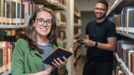 Em uma biblioteca iluminada, com prateleiras cheias de livros antigos dispostos lateralmente ao longo de um corredor estreito, uma mulher de pele branca, ocupa o centro da imagem. Ela está de pé, sorrindo diretamente para a câmera, usando óculos de armação preta, blazer verde e blusa cinza. Segura um livro aberto, de capa escura, próximo ao peito. Seus cabelos castanhos, lisos e longos, estão soltos. Ao fundo, levemente desfocado, um homem de pele preta sorri enquanto segura um livro; ele veste camiseta preta e calça escura. Ambos estão entre as estantes repletas de livros.