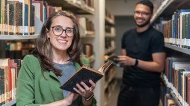 Em uma biblioteca iluminada, com prateleiras cheias de livros antigos dispostos lateralmente ao longo de um corredor estreito, uma mulher de pele branca, ocupa o centro da imagem. Ela está de pé, sorrindo diretamente para a câmera, usando óculos de armação preta, blazer verde e blusa cinza. Segura um livro aberto, de capa escura, próximo ao peito. Seus cabelos castanhos, lisos e longos, estão soltos. Ao fundo, levemente desfocado, um homem de pele preta sorri enquanto segura um livro; ele veste camiseta preta e calça escura. Ambos estão entre as estantes repletas de livros.