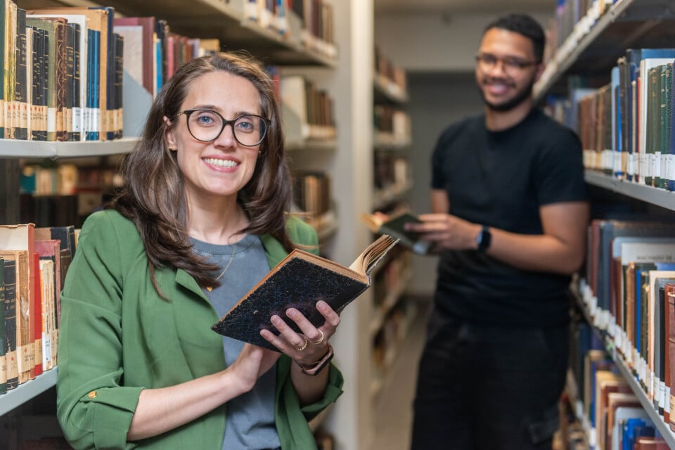 Em uma biblioteca iluminada, com prateleiras cheias de livros antigos dispostos lateralmente ao longo de um corredor estreito, uma mulher de pele branca, ocupa o centro da imagem. Ela está de pé, sorrindo diretamente para a câmera, usando óculos de armação preta, blazer verde e blusa cinza. Segura um livro aberto, de capa escura, próximo ao peito. Seus cabelos castanhos, lisos e longos, estão soltos. Ao fundo, levemente desfocado, um homem de pele preta sorri enquanto segura um livro; ele veste camiseta preta e calça escura. Ambos estão entre as estantes repletas de livros.