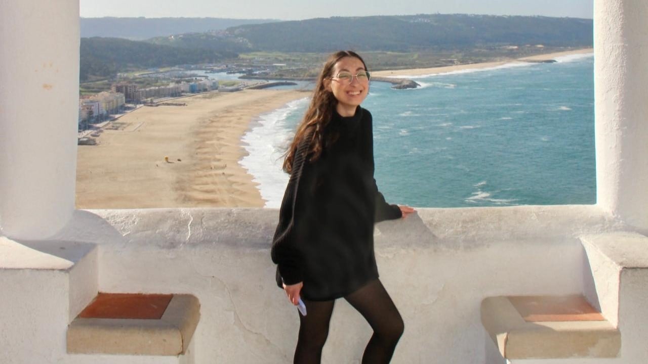 Ao fundo da imagem, vê-se uma ampla praia de areia dourada que se estende em curva pela costa, à esquerda, e um mar azul à direita, com pequenas ondas quebrando próximas à areia. No horizonte, há uma área de morros verdes e edificações urbanas ao longe. A foto foi tirada de um ponto elevado, em uma estrutura branca com colunas e bancos de alvenaria integrados. No centro da imagem, de pé em frente a um parapeito, está Tiele Pietra Alves, uma mulher branca, de cabelos longos e castanhos, soltos. Ela sorri, usando óculos de armação clara, uma blusa preta comprida e meia-calça preta. A mão esquerda está apoiada no parapeito e a direita segura um objeto claro, possivelmente uma máscara.