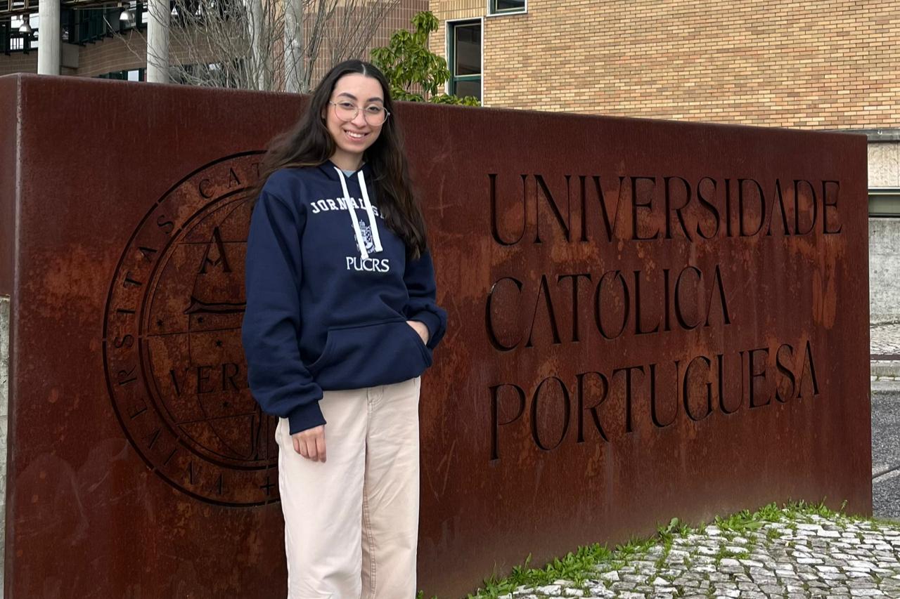 Em primeiro plano, à direita da imagem, há uma placa grande de metal oxidado com a inscrição "Universidade Católica Portuguesa" em letras recortadas e, à esquerda, o brasão da instituição. Ao fundo, observa-se parte do prédio da universidade, com fachada de tijolos aparentes e janelas retangulares, além de uma árvore sem folhas e uma pequena área verde com arbustos. Ao centro da imagem, está Tiele Pietras Alves, uma jovem de pele branca, cabelos longos e lisos, castanhos escuros. Ela usa óculos de grau com armação redonda, veste um moletom azul-marinho com a inscrição “Jornalismo PUCRS” em branco, e calça bege clara. O chão é composto por pedras portuguesas em tons de cinza e branco, com um trecho de grama verde ao lado da placa.