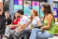 Cena em área externa da PUCRS, com seis jovens sentados em um banco de concreto, em frente a uma parede com painéis coloridos de divulgação institucional. Os cartazes ao fundo exibem mensagens sobre inovação, laboratórios e criatividade, com elementos gráficos em tons de azul, rosa, roxo e laranja.