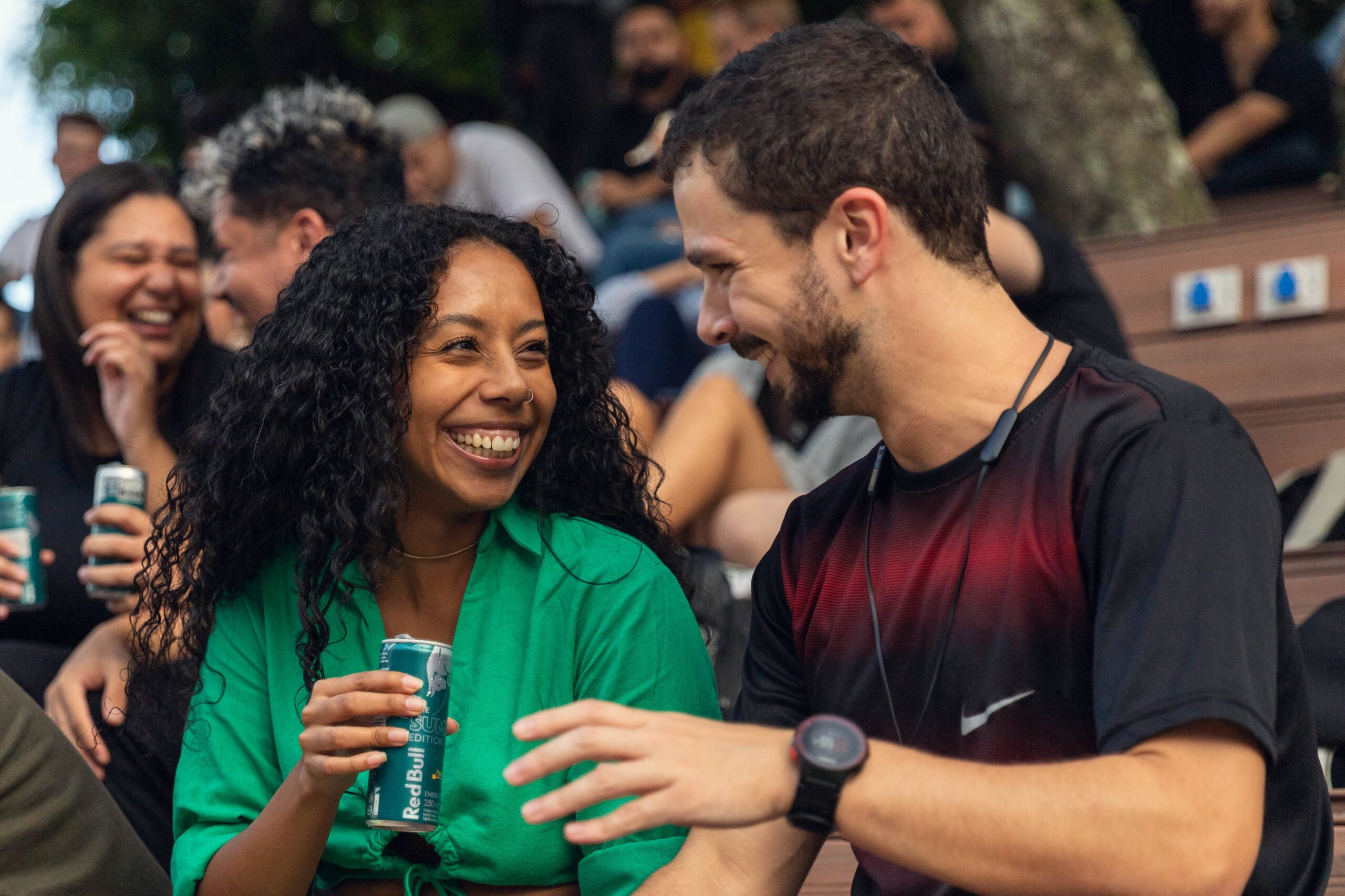 Em um espaço ao ar livre, estudantes estão sentados em arquibancadas de madeira, sob a sombra de árvores. Em destaque no centro da imagem, uma jovem de cabelos cacheados e blusa verde sorri amplamente enquanto conversa com um colega ao lado, que veste camiseta preta com vermelho e fones de ouvido pendurados no pescoço. Ambos seguram latas de bebida e trocam olhares alegres. Ao fundo, outros jovens também riem e conversam, criando um clima descontraído e acolhedor de convivência universitária.